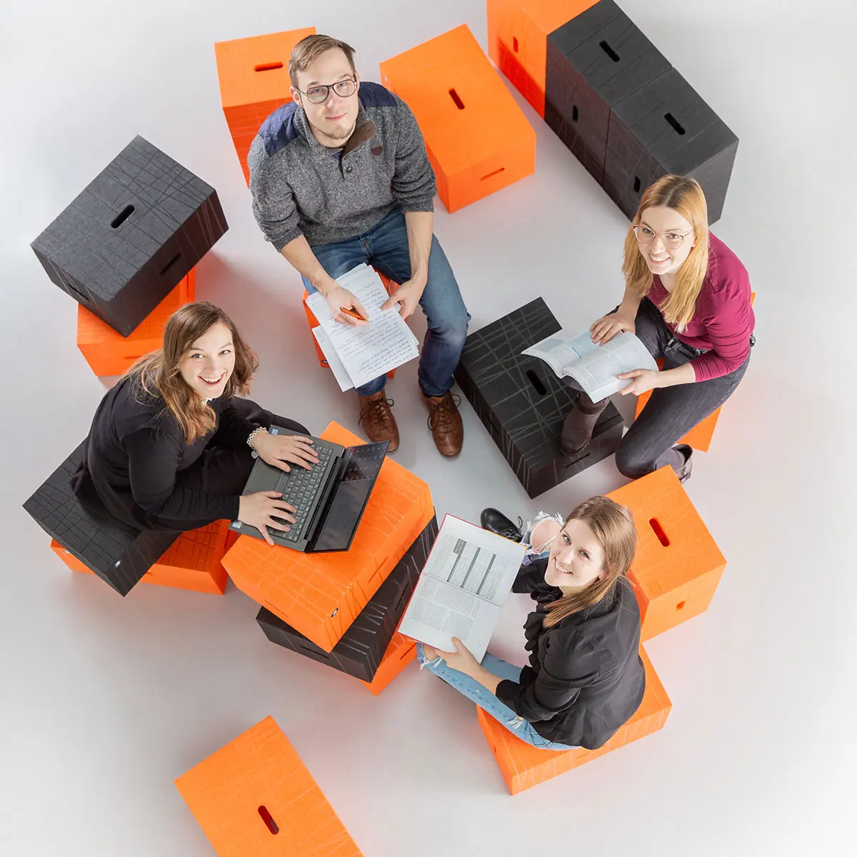 Studierende Crossmedia-Redaktion/Public Relations sitzen auf Sitzwürfeln und lachen in die Kamera.