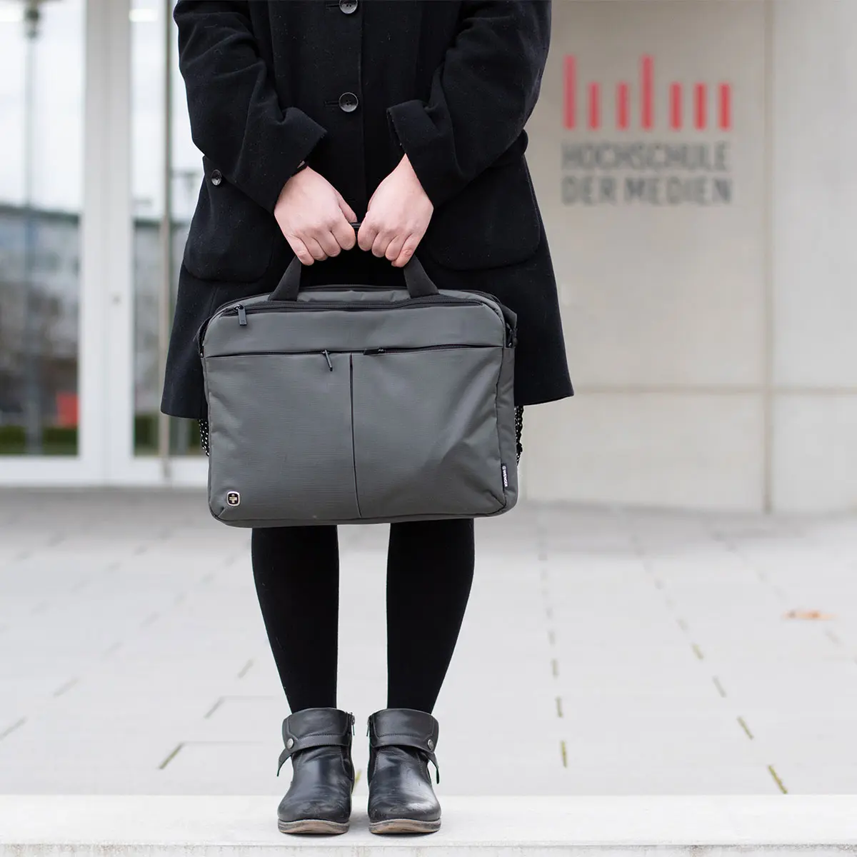 Studentin Crossmedia-Redaktion/Public Relations steht mit Laptoptasche vor dem HdM Gebäude. Zu sehen ist das HdM Logo am Gebäude im Hintergrund.