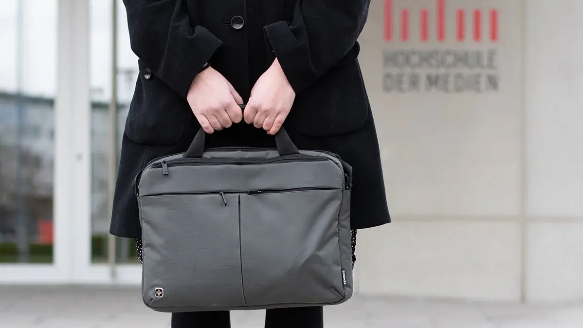 Studentin Crossmedia-Redaktion/Public Relations steht mit Laptoptasche vor dem HdM Gebäude. Zu sehen ist das HdM Logo am Gebäude im Hintergrund.