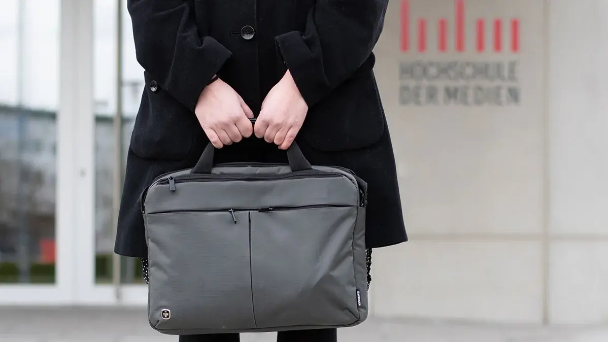 Studentin Crossmedia-Redaktion/Public Relations steht vor dem Gebäude der HdM und hält eine Laptoptasche vor sich.