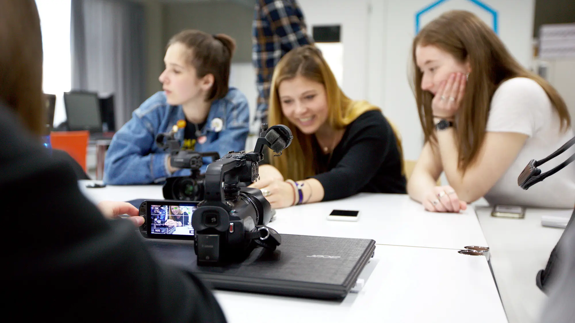 Studierende Crossmedia-Redaktion/Public Relations sitzen am Redaktionstisch im edit.Lab, der Lehrredaktion des Studiengangs, und besprechen sich. Im Vordergrund ist eine Videokamera und ein Laptop zu sehen.